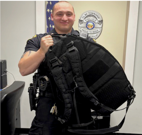 Police officer in uniform holding a black backpack with the Clay Township Police badge in the background.