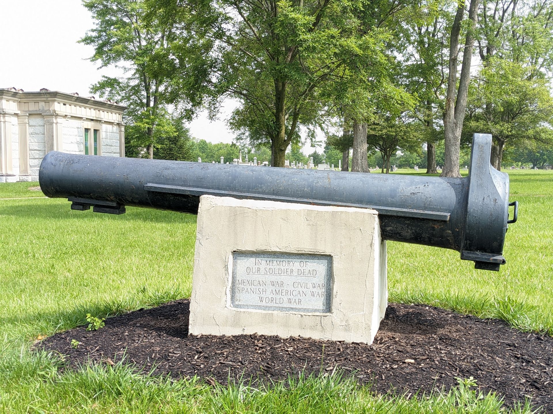 Cannon on a concrete base in a grassy area with trees and a building in the background.