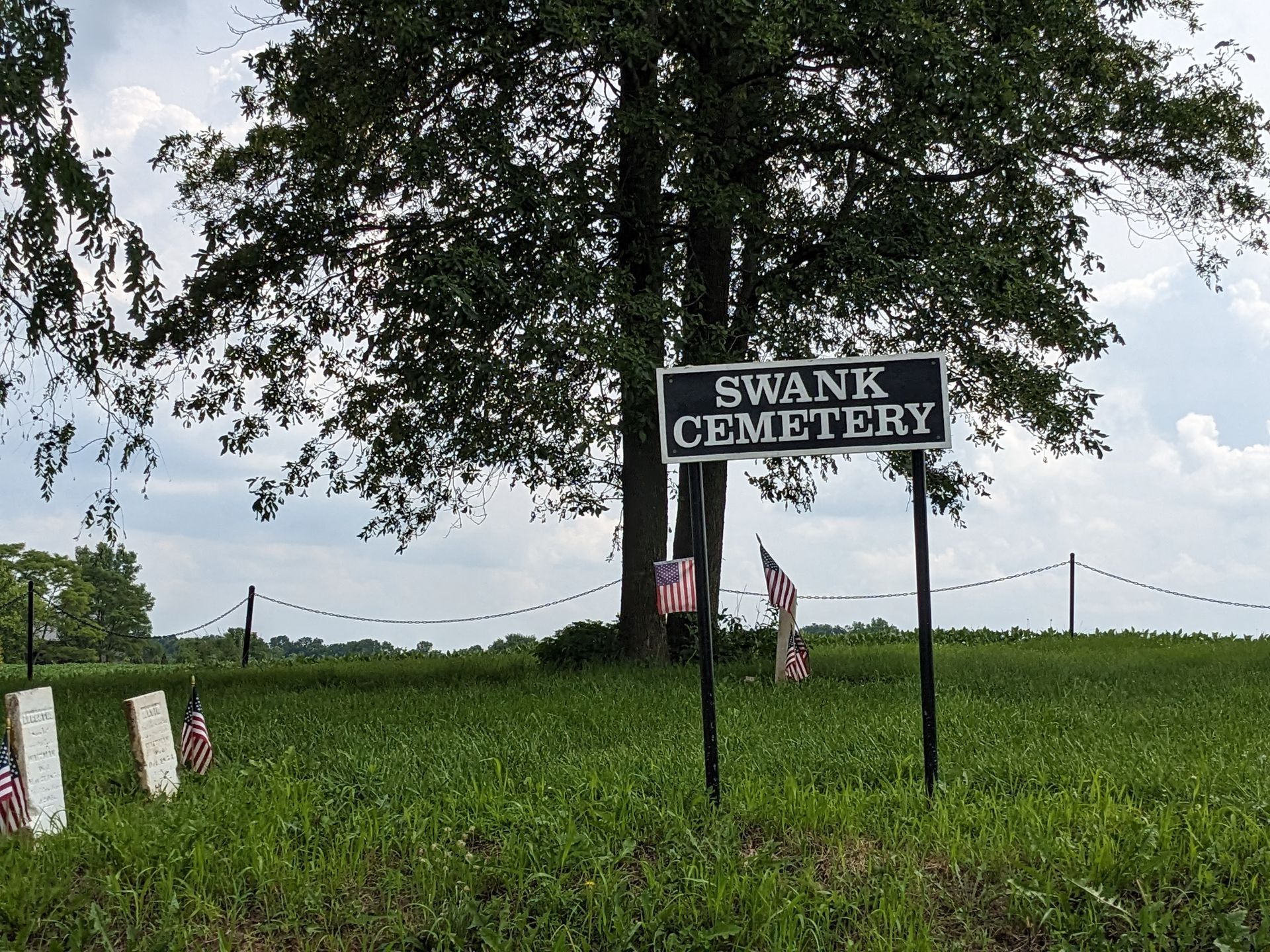 Sign for Swank Cemetery; headstones and American flags in a field under a cloudy sky.