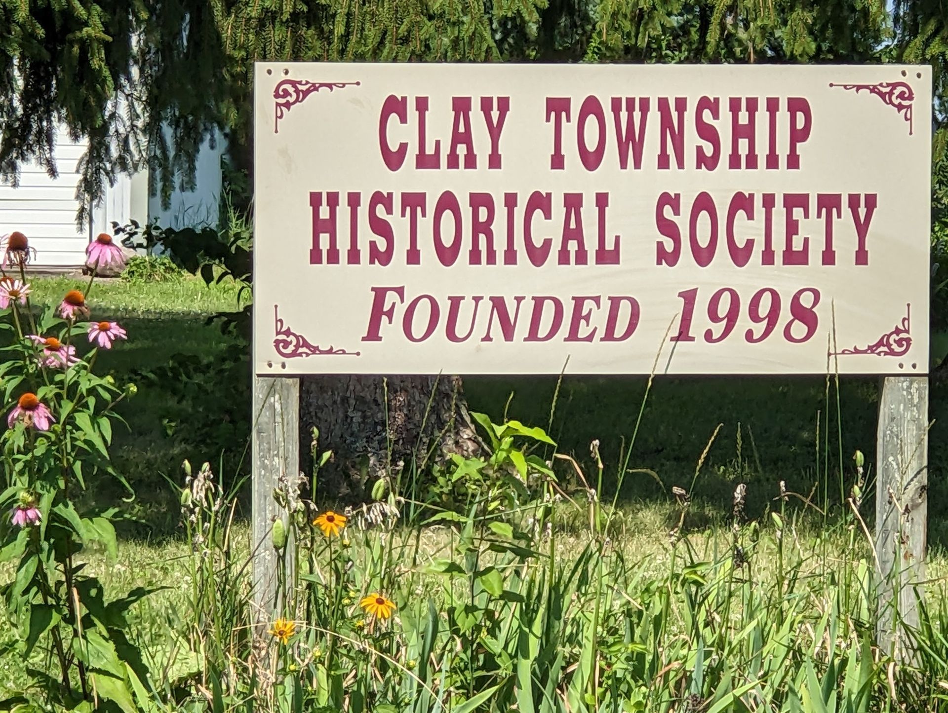 Sign for Clay Township Historical Society, founded 1998, in a grassy setting with flowers and a white building in the background.