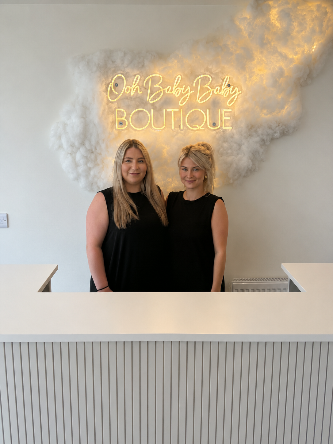 Two women standing behind a white reception desk under a glowing “Oh Baby Boutique” sign