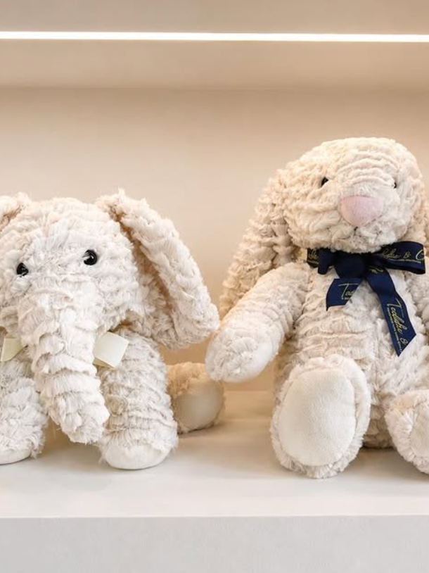 Two white crocheted plush animals sitting on a shelf, one with a black ribbon and pink nose