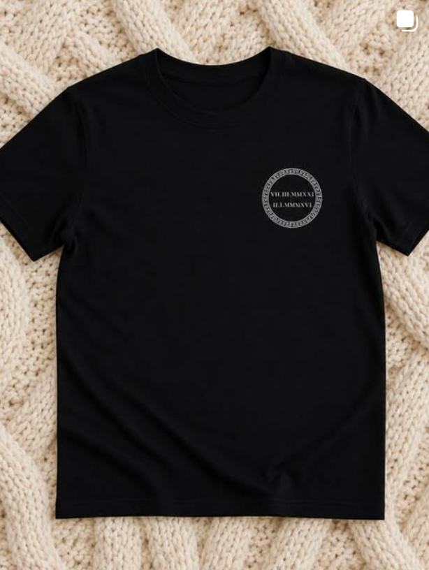 Black T-shirt with a small circular logo on the left chest, laid flat on a beige textured background