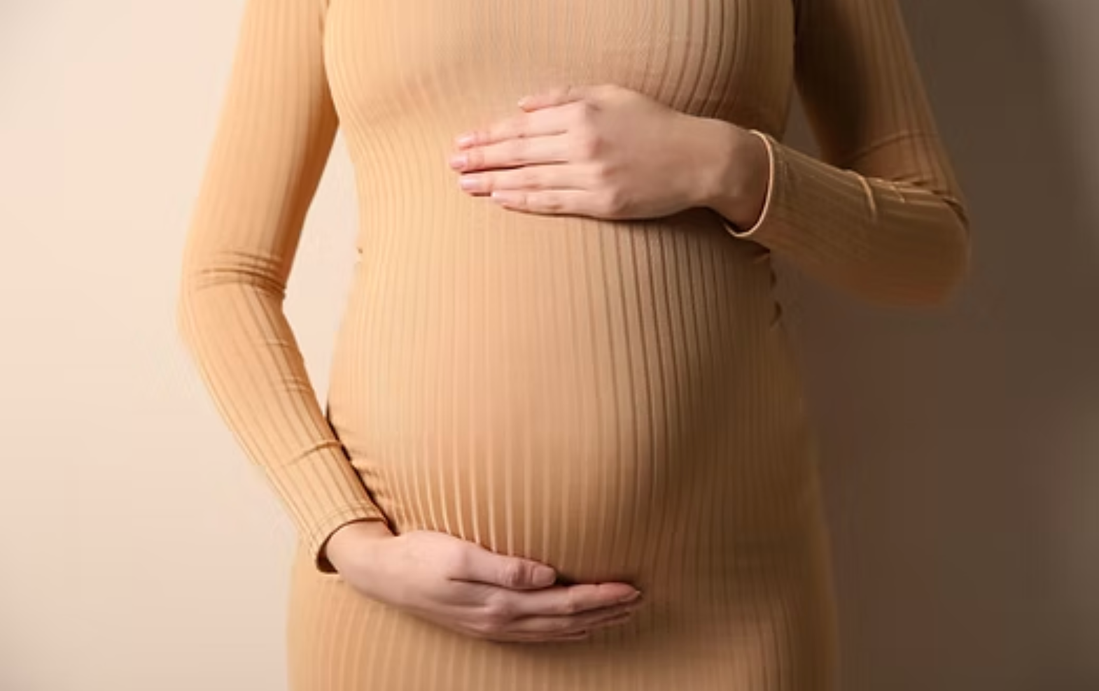Pregnant person in a beige ribbed dress gently holding their belly
