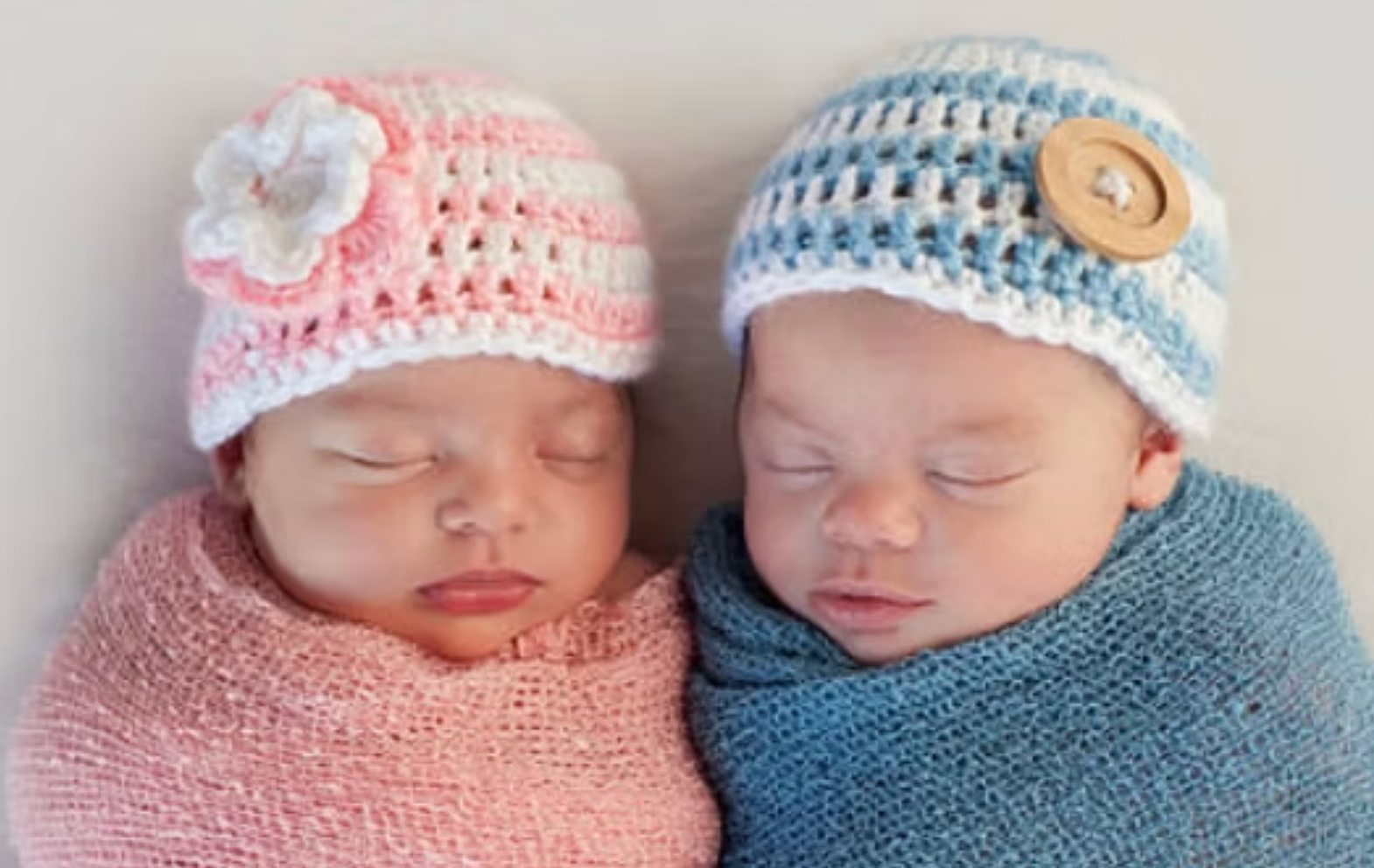Two sleeping newborns swaddled in pink and blue crochet hats and blankets