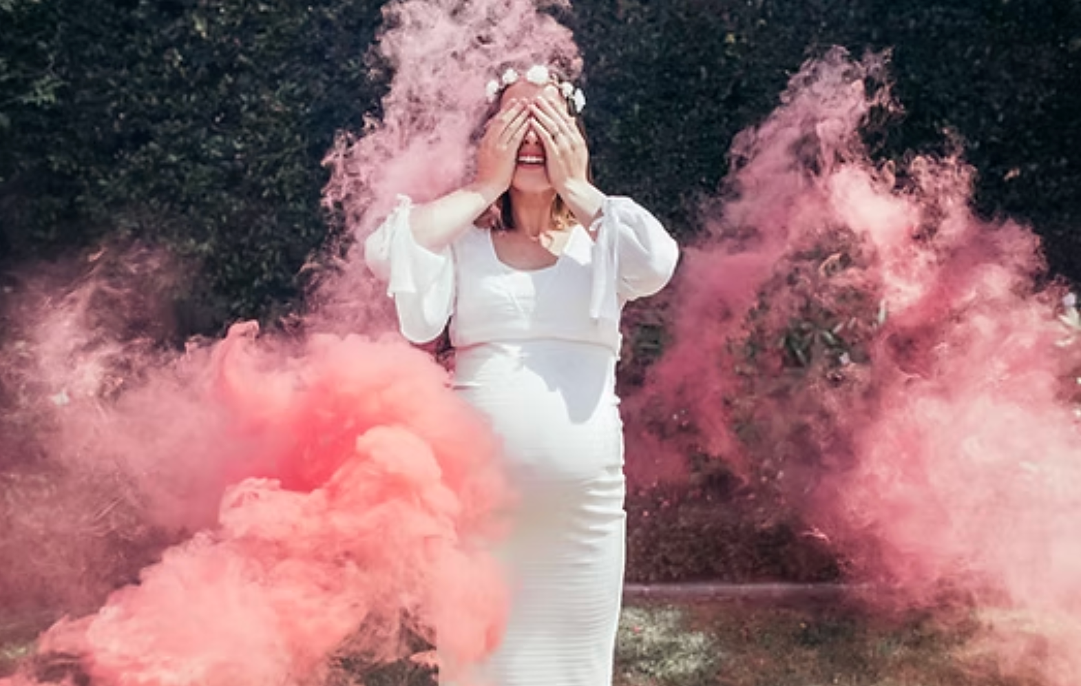 Woman in a white dress standing in pink smoke outdoors, covering her face with her hands.