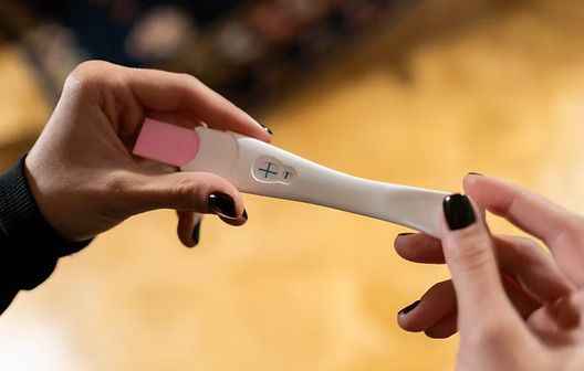 Hands holding a positive pregnancy test with a pink cap in a warm indoor setting