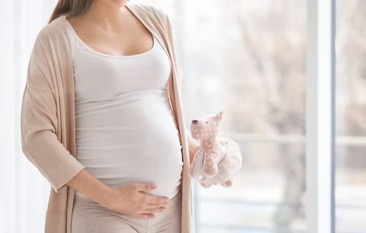 Pregnant person in a beige cardigan holding her belly by a bright window