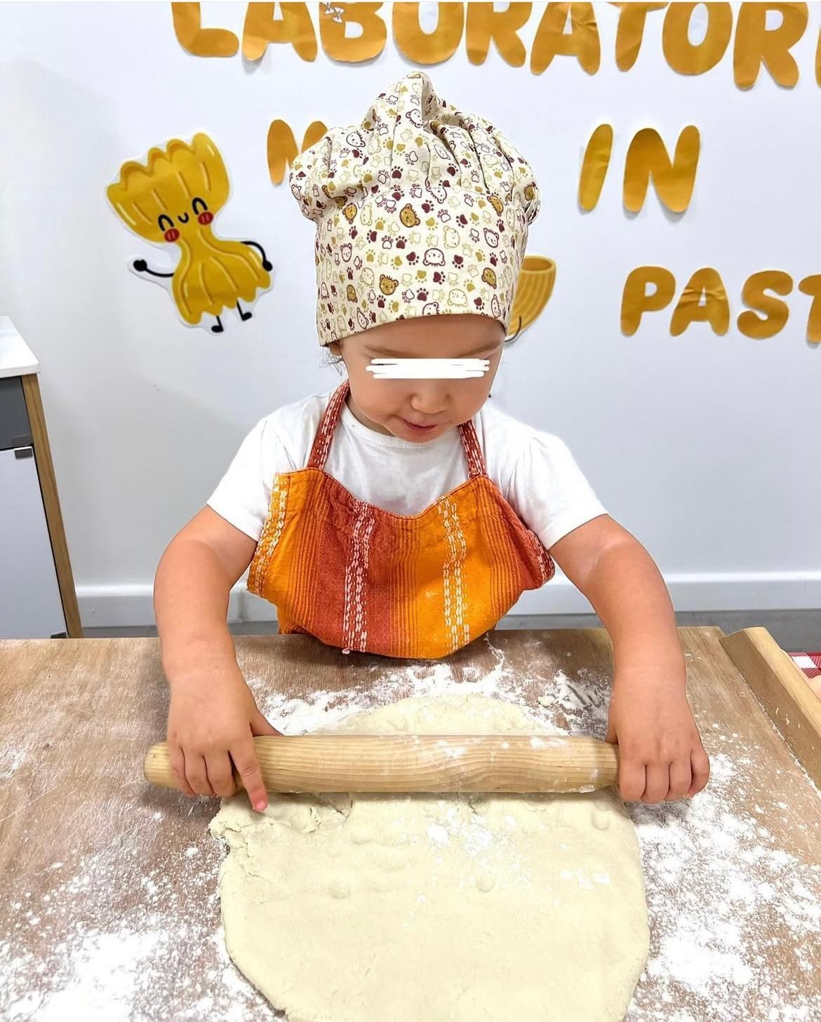 Un ragazzino che indossa un cappello da cuoco e un grembiule sta stendendo la pasta con un mattarello
