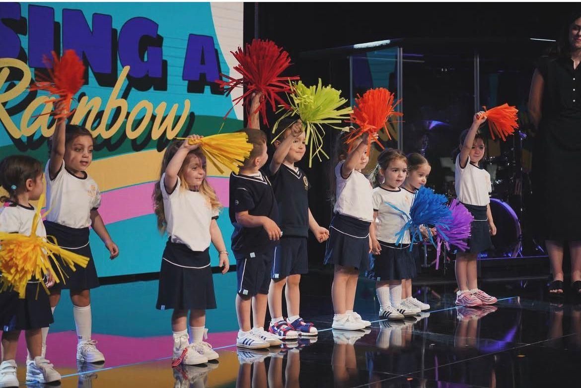 Un gruppo di bambini tiene in mano dei pompon davanti a un cartello che dice canta un arcobaleno