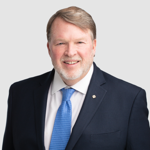 Bruce Stubbs in a blue suit smiles at the camera against a gray background.
