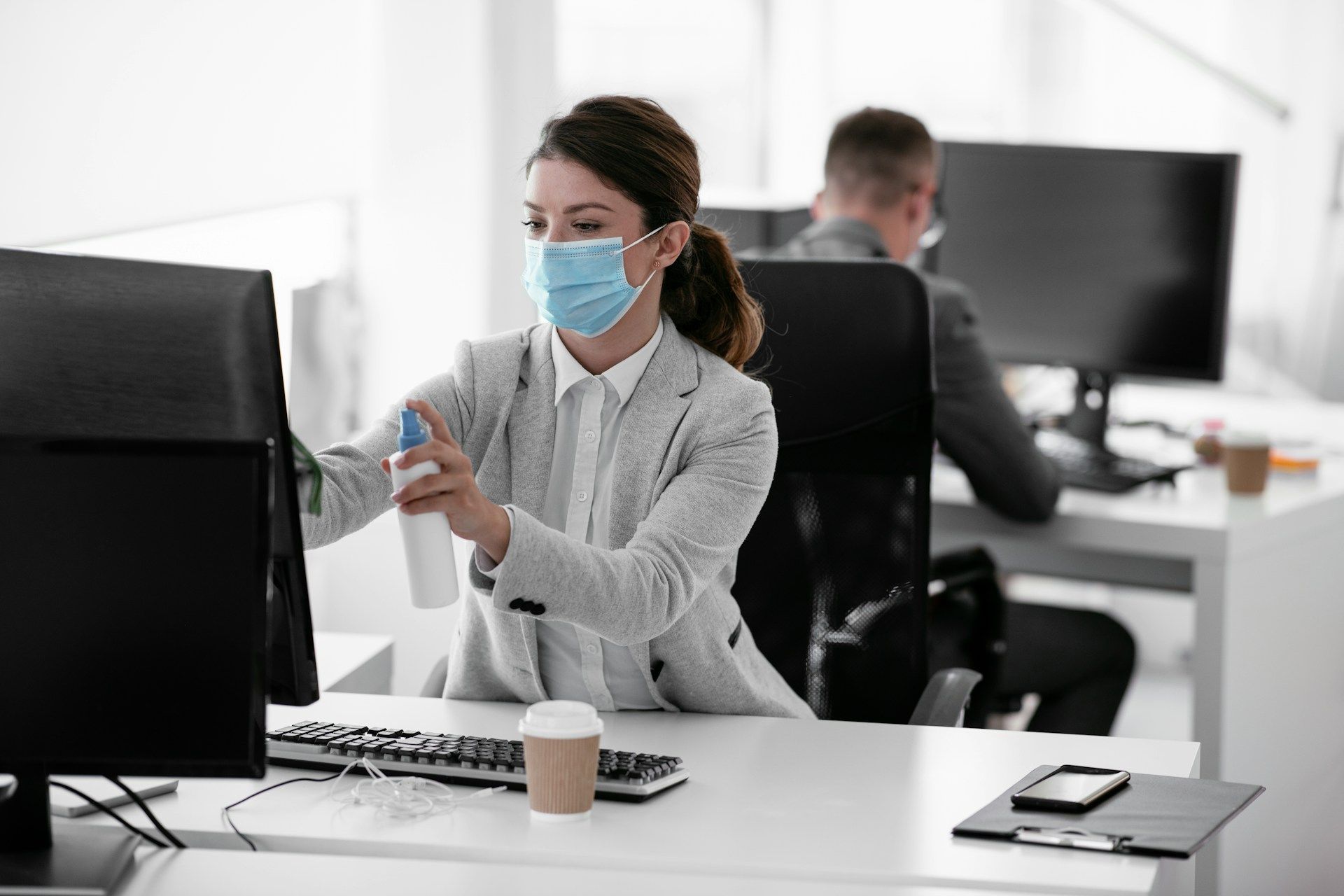 Woman with respiratory mask cleaning her computer monitor in a brightly lit office
