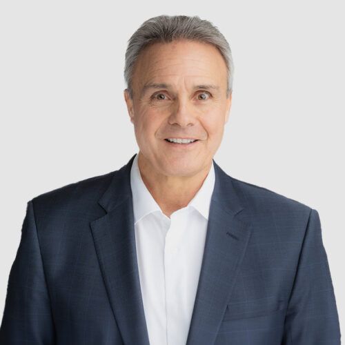 Mark Churella in a suit smiles, white shirt, grey hair, studio shot.