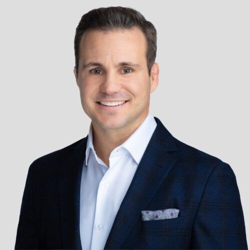 Ryan Churella in a navy blazer and white shirt, smiling. Gray background, pocket square.