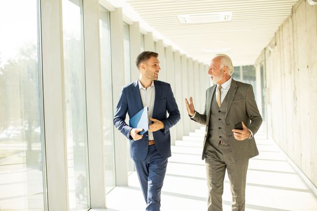 Two men in suits walking and talking in a well-lit hallway with large windows.
