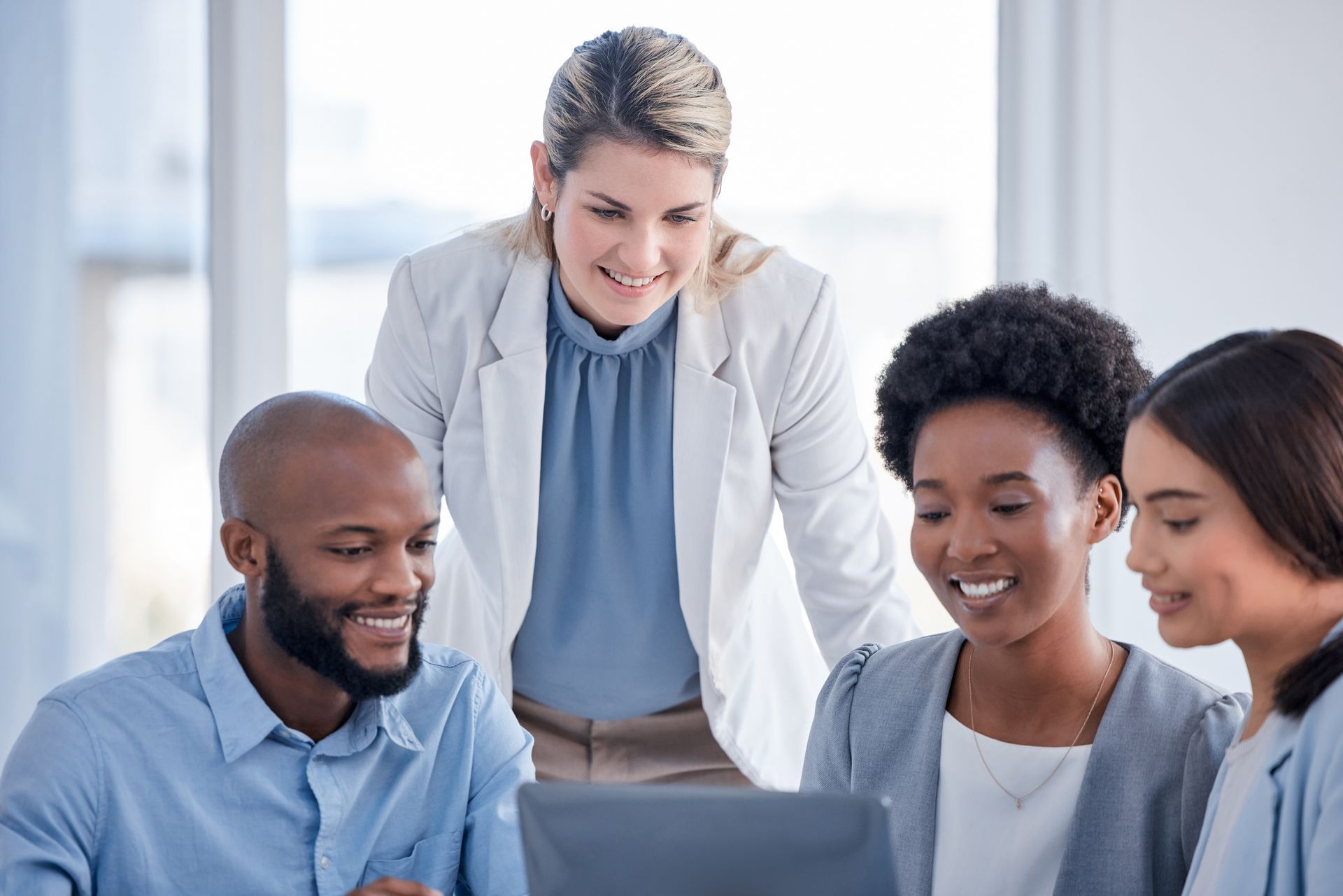 Four people, one standing, looking at a laptop in a bright office.