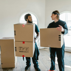 Two people carrying cardboard boxes in a bright empty room