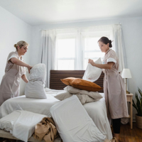 Two people making a bed in a bright bedroom with white bedding and curtains