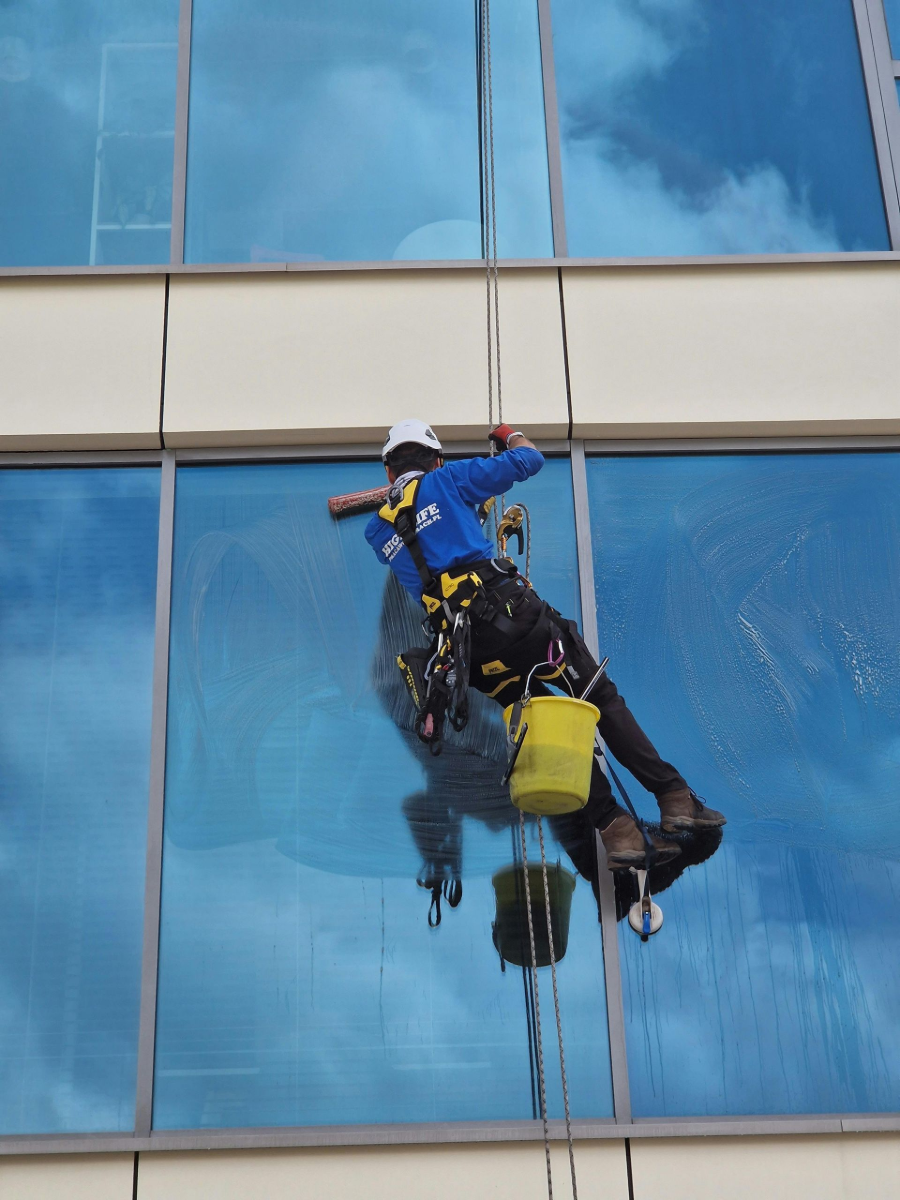 Rope-access worker in blue gear suspended on a glass building facade, cleaning windows
