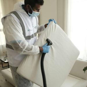 Person in protective gear sanitizing a white cushion with a spray hose indoors