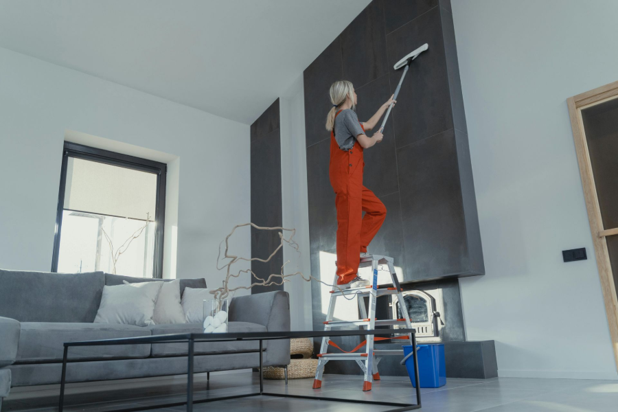 Person in orange overalls painting a tall dark wall from a ladder in a bright living room
