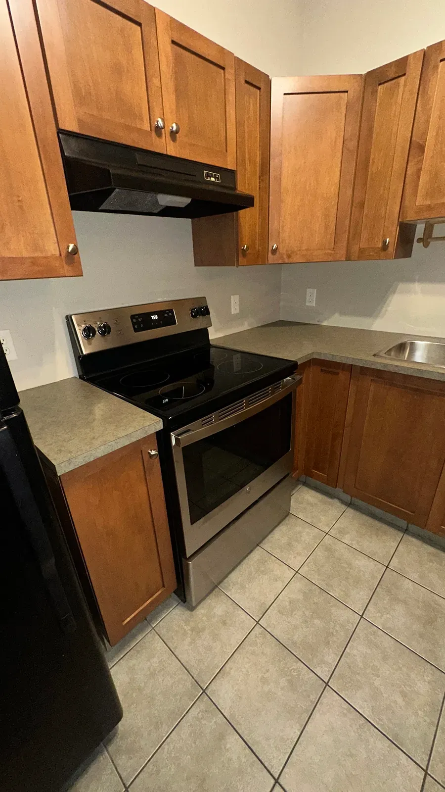 Compact kitchen with wooden cabinets, black stove, stainless sink, and beige tiled floor.