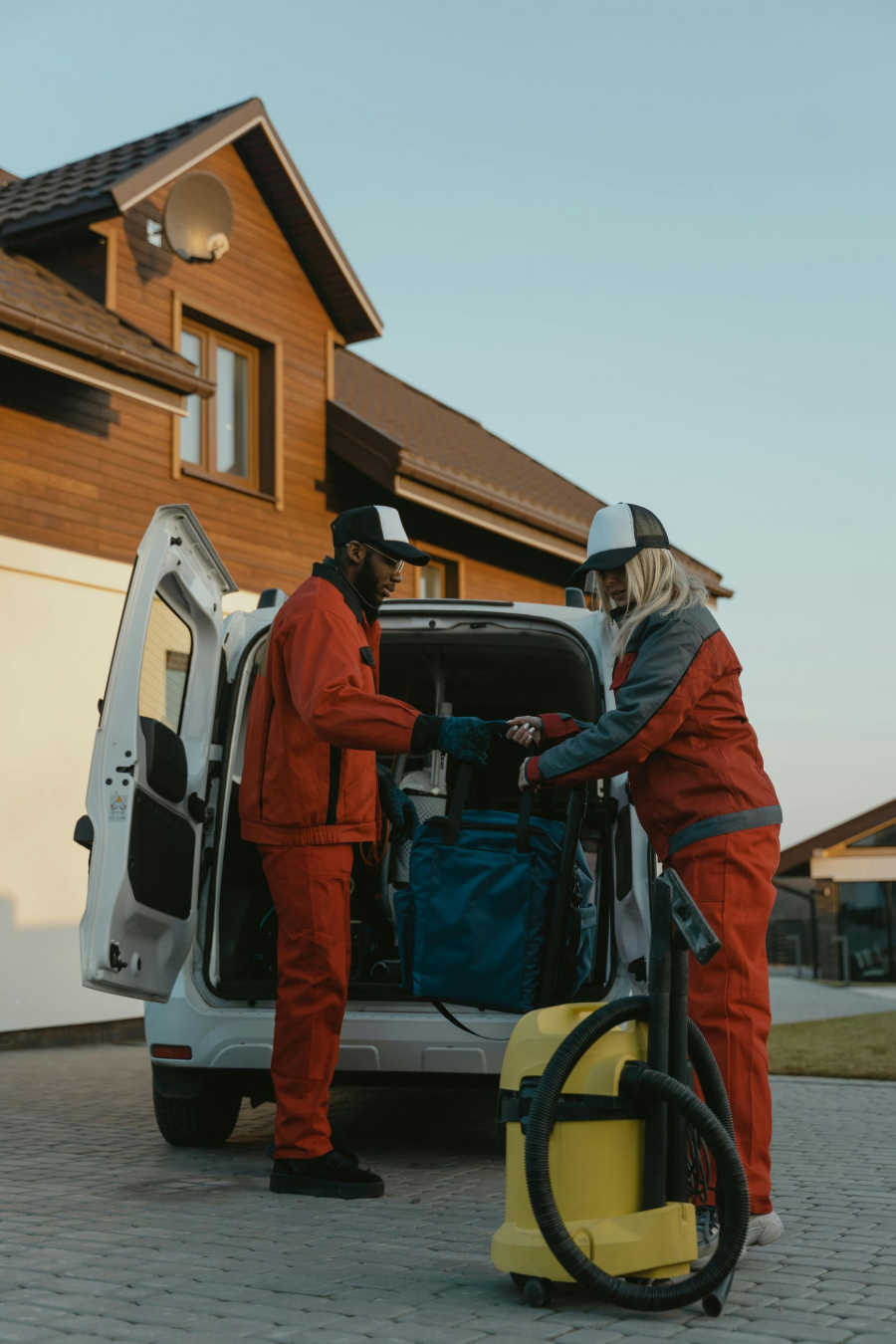 Two workers in orange uniforms loading equipment into a white van outside a house