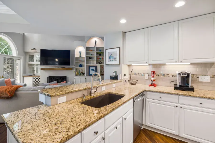 Bright kitchen with white cabinets, granite island, stainless sink, and built-in fireplace in background