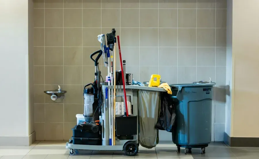 Cleaning cart and supplies beside a bathroom sink and trash bin in a tiled restroom