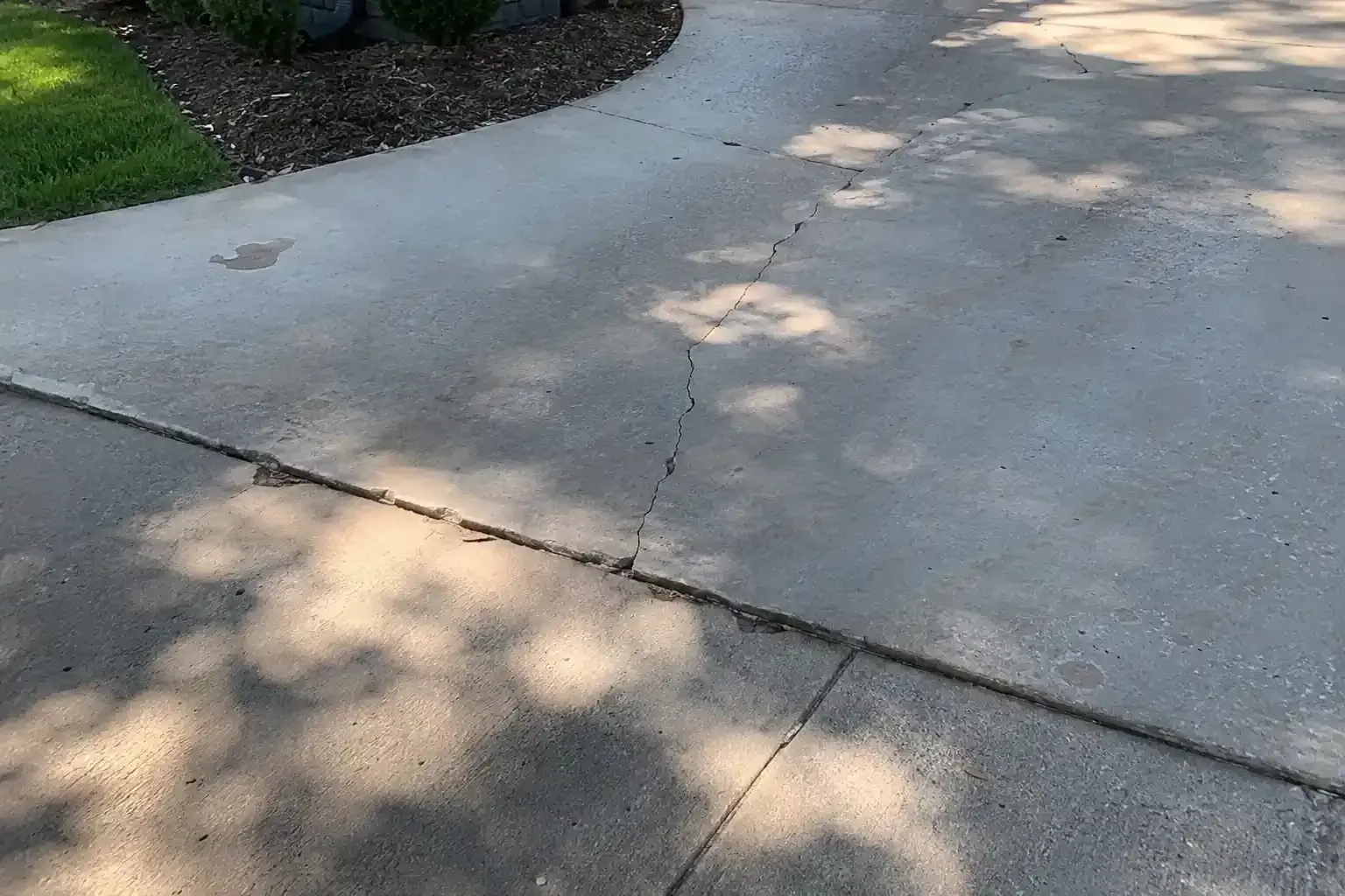 Cracked concrete driveway bordered by grass and mulch, with dappled tree shadows.