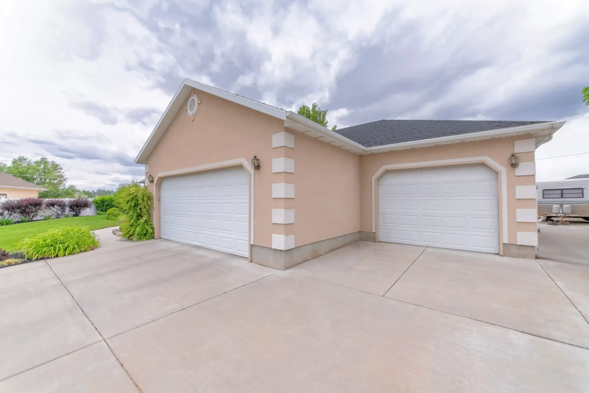 Two-car beige garage with white doors with a new wide concrete driveway under a cloudy sky