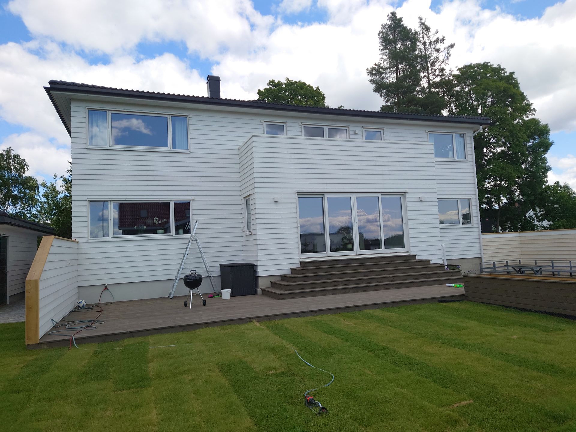 Hvitt toetasjes hus med flere vinduer, en stor glassdør og en terrasse på en grønn plen under en overskyet himmel.