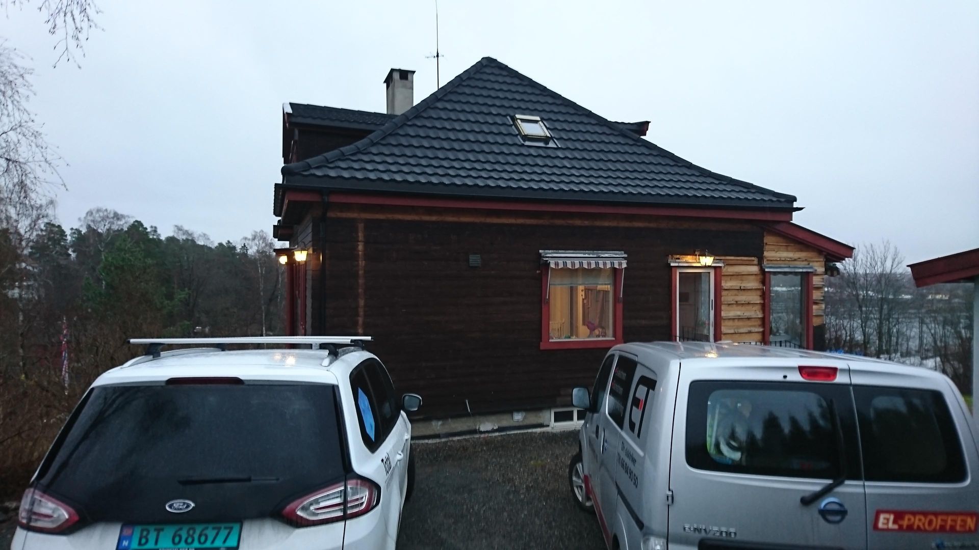 Mørkebrunt trehus med mørkt tegltak, parkerte biler foran, overskyet himmel.