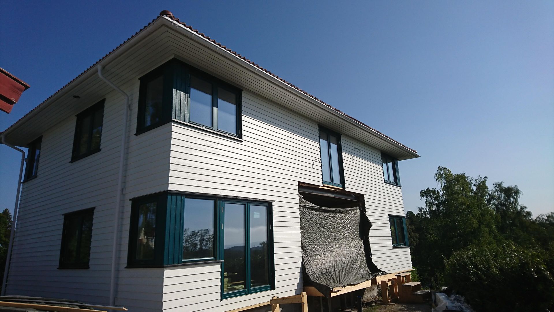 To-etasjes hvitt hus med mørkeblå vindusrammer, under en knallblå himmel. Byggingen er i gang på høyre side.