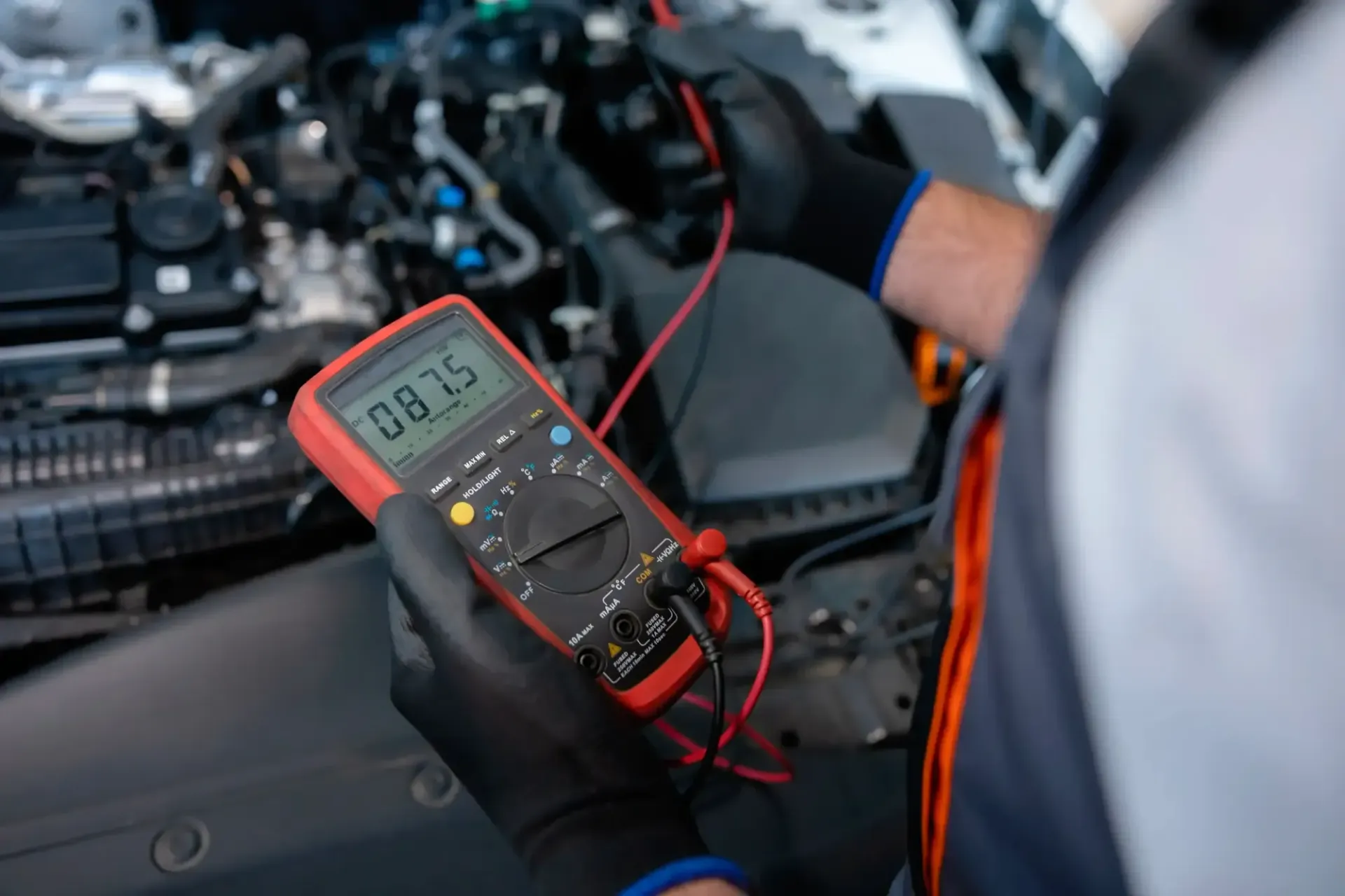 Mechanic using a red multimeter to test car engine wiring under the hood