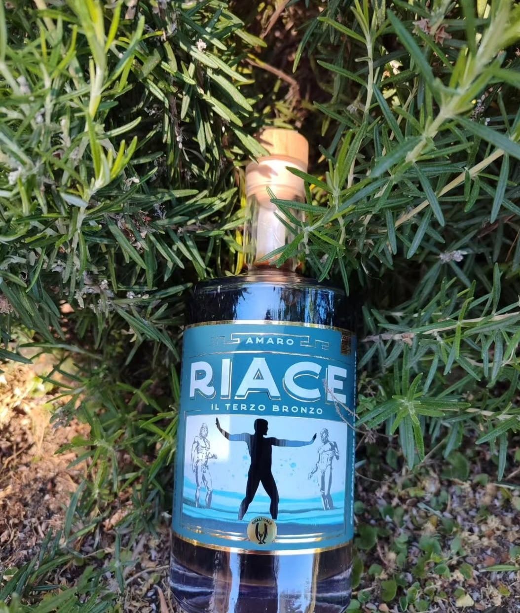 Bottiglia blu di liquore con l’etichetta «RIACE», immersa tra arbusti verdi, con una scena di spiaggia sull’etichetta.