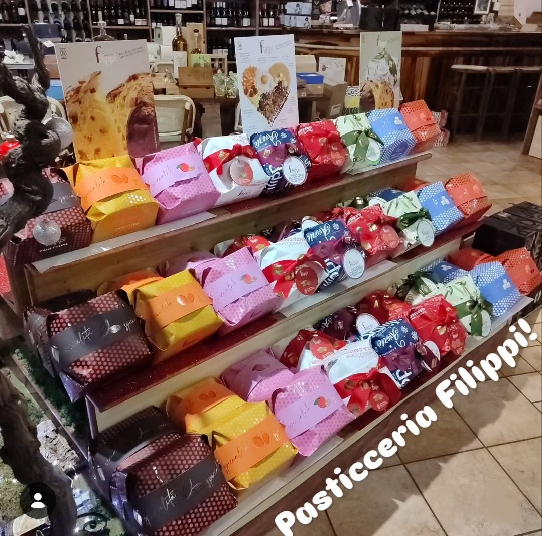 File di cesti regalo colorati e confezionati, disposti su scaffali di legno in un negozio, con la scritta “Pasticceria” visibile.