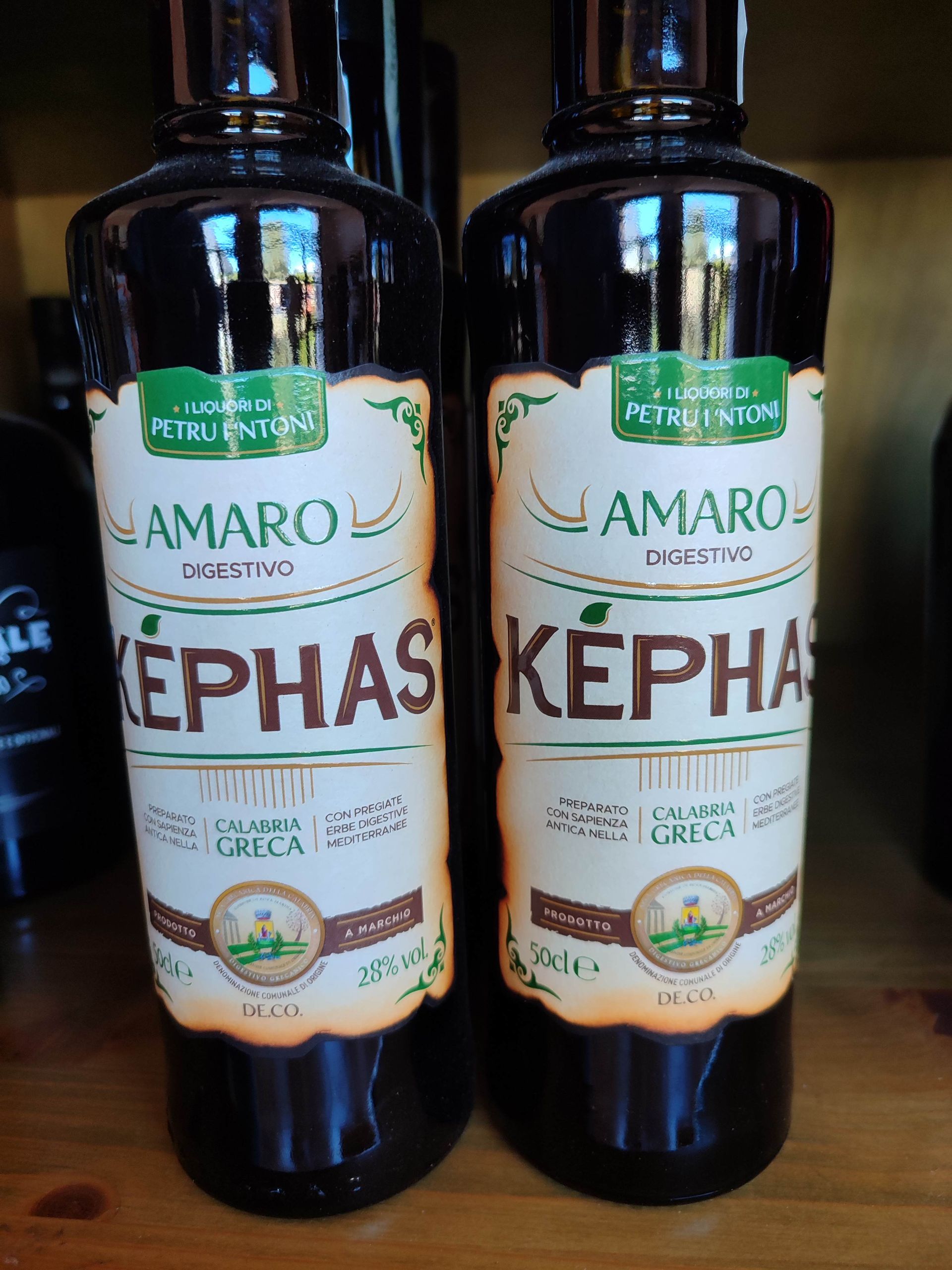 Due bottiglie scure di AMARO KEPHAS con etichette beige e tappi verdi su uno scaffale.