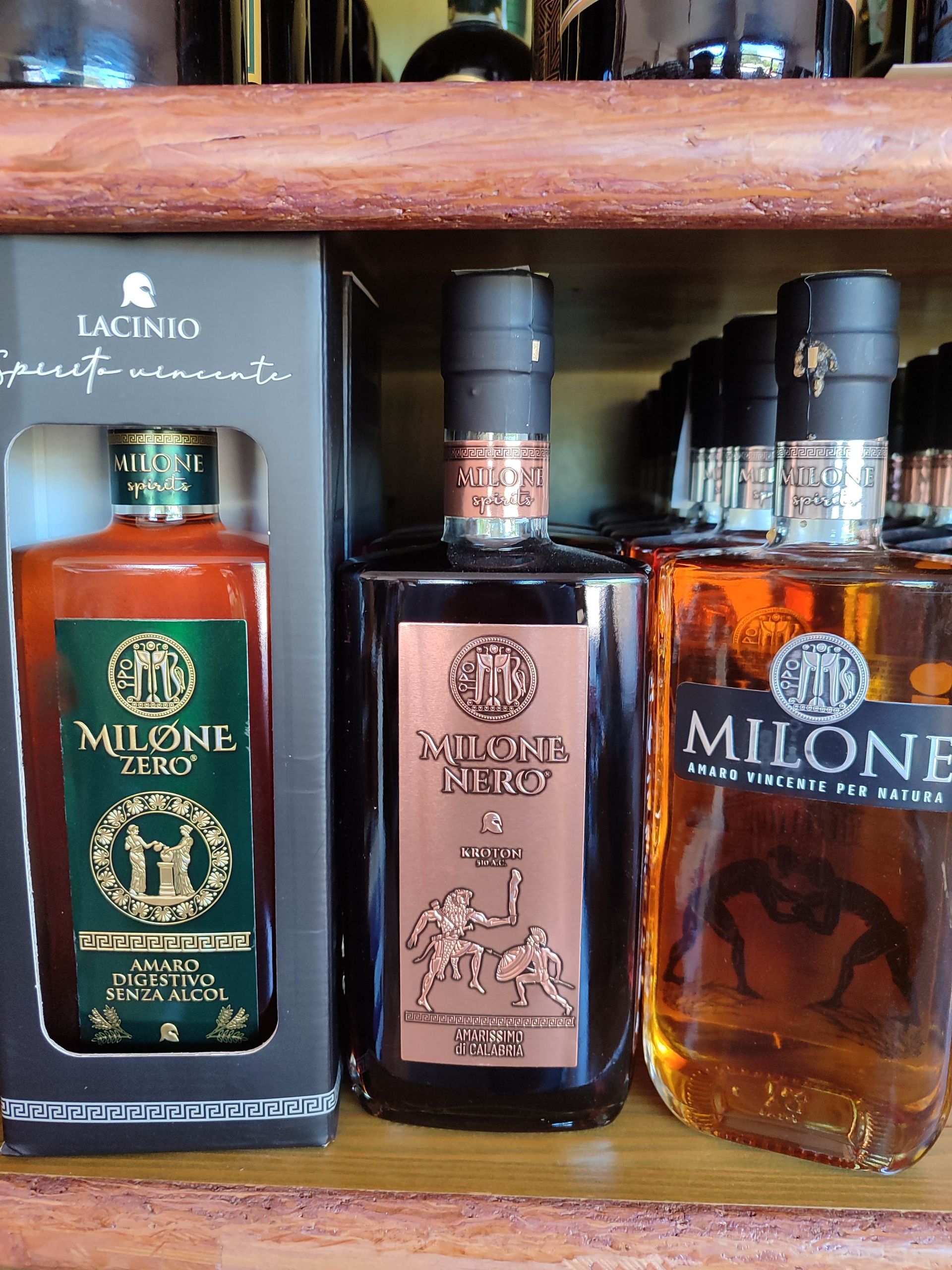 Tre bottiglie di liquore Milone esposte su una mensola di legno, una in una scatola a sinistra.