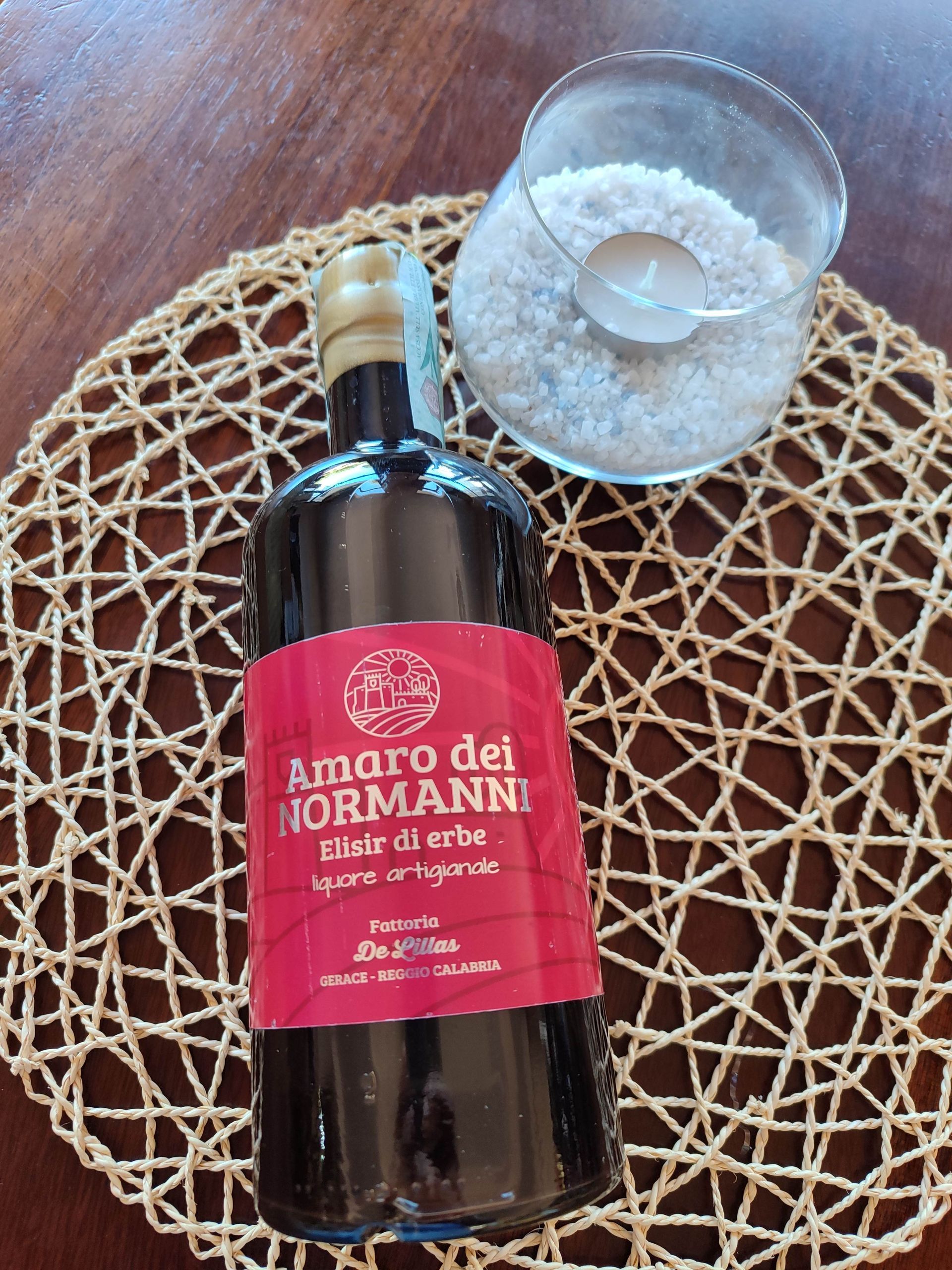 Bottiglia di Amaro dei Normanni con etichetta rossa accanto a un bicchiere con ghiaccio, su una tovaglietta intrecciata appoggiata a un tavolo di legno
