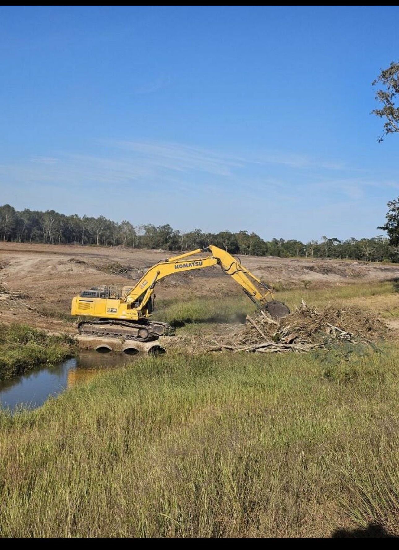Yellow excavator digging dirt beside a small water channel in a grassy field under a clear blue sky