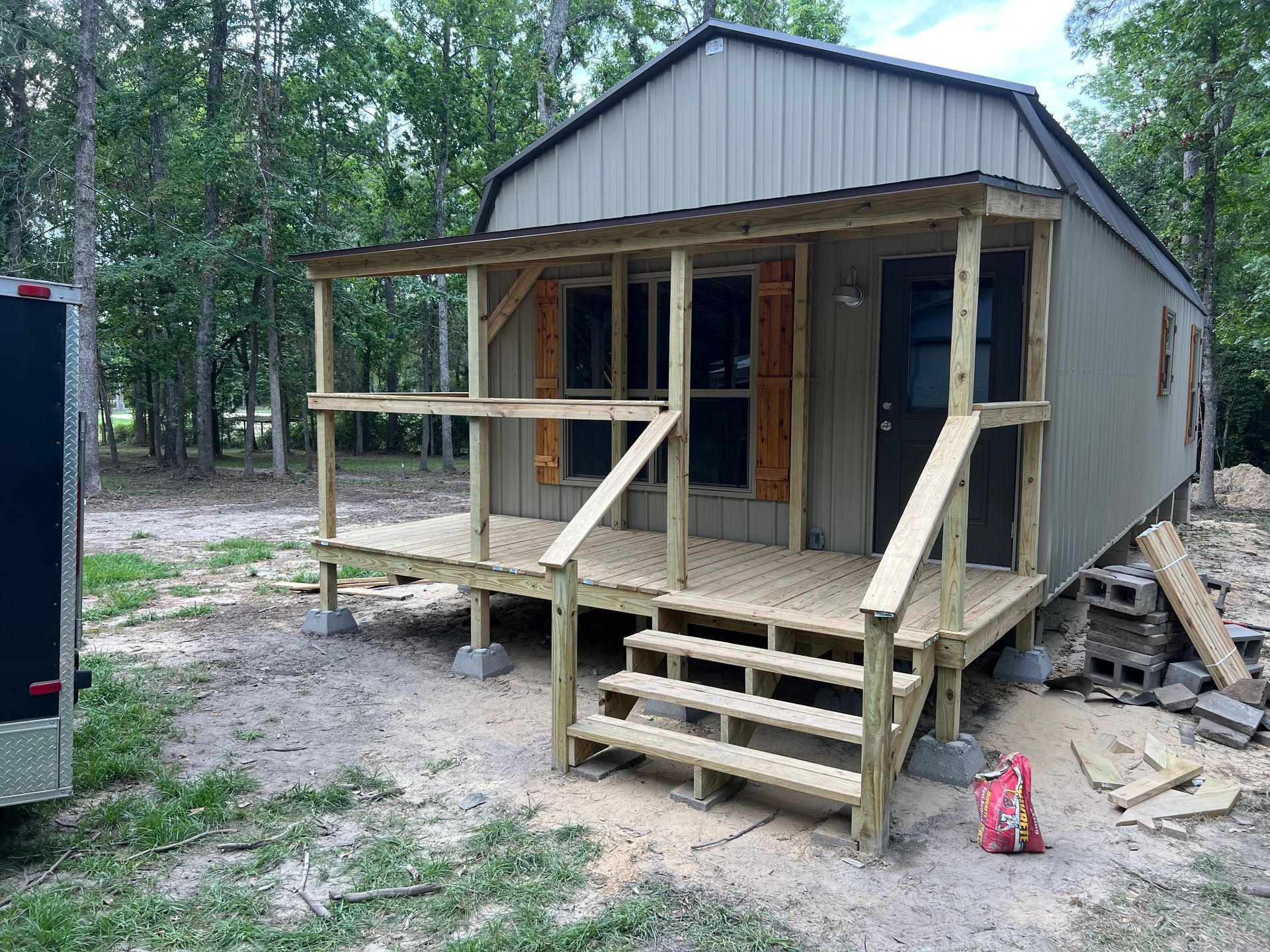 Small gray cabin under construction with unfinished porch, steps, and support posts in a wooded area