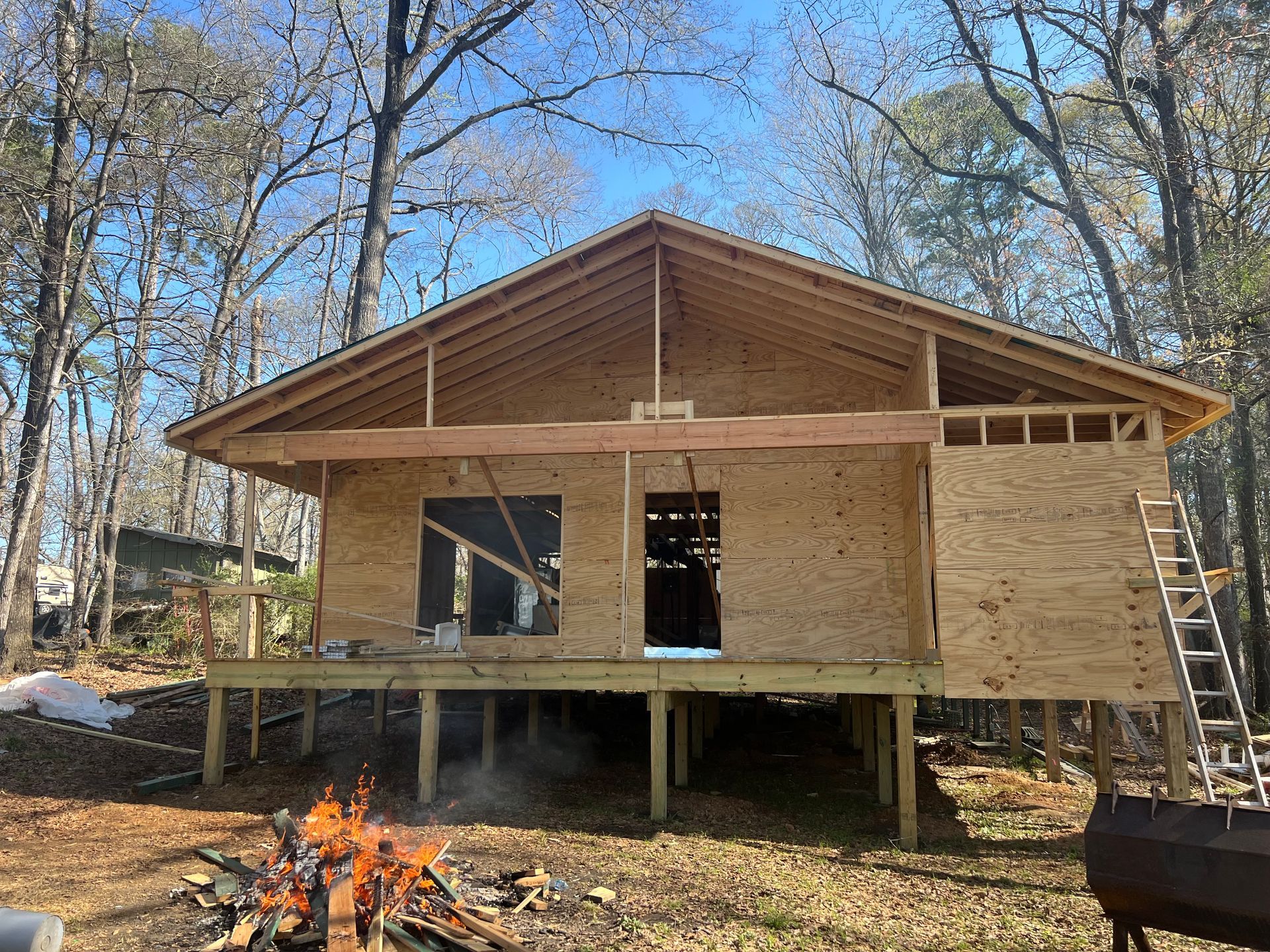 Partially built wooden cabin with porch and ladder in a wooded yard, with a small fire in the foreground