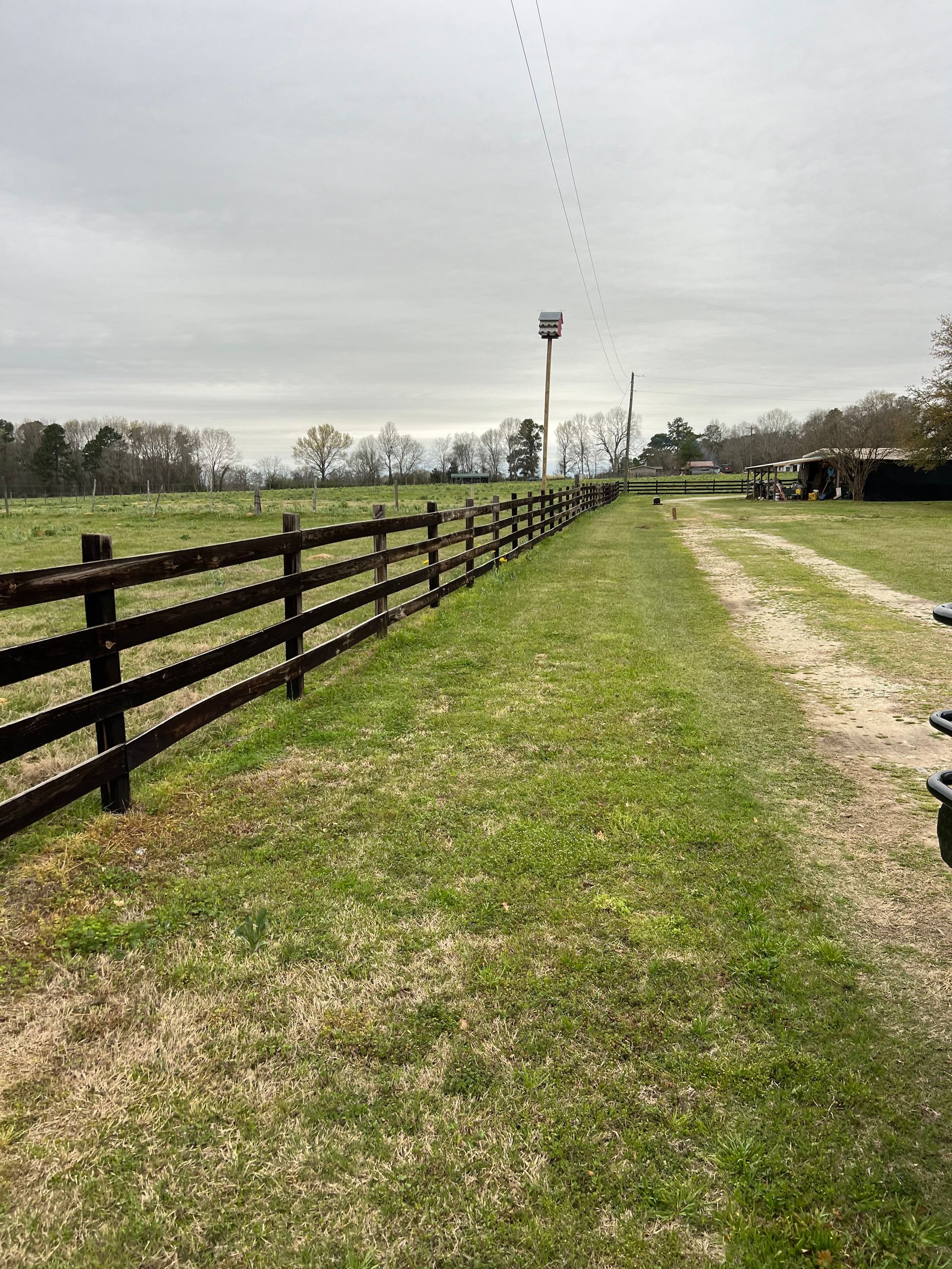 Grassy farm lane beside a dark wooden fence, leading to a distant barn under a cloudy sky.