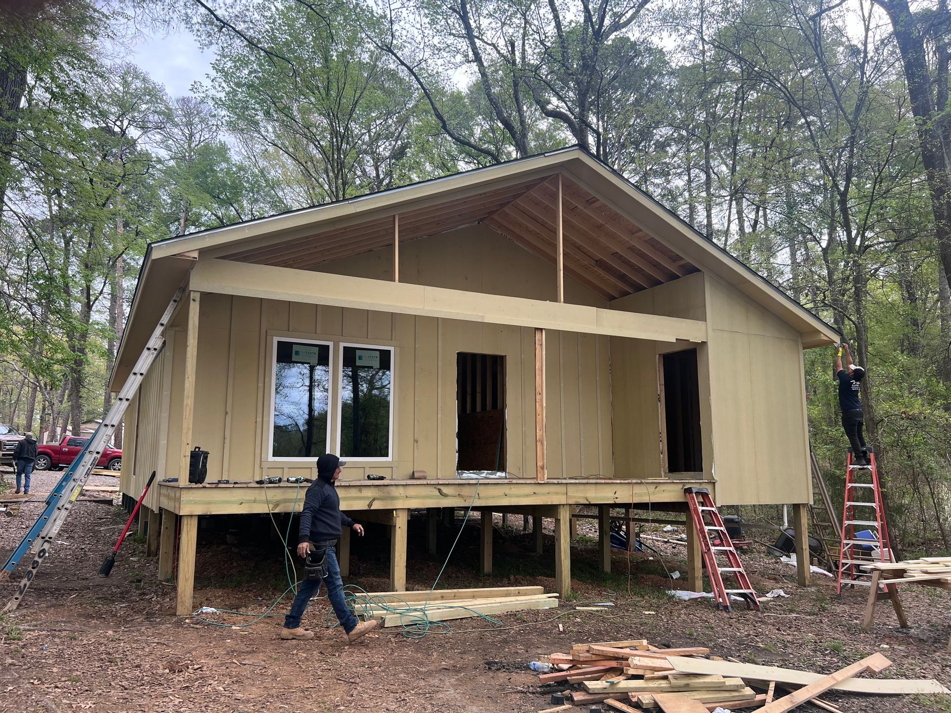 Partially built tan cabin with workers on ladders in a wooded area