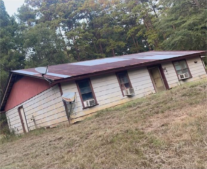 Tilted weathered house with a rusted metal roof in a grassy, wooded area.