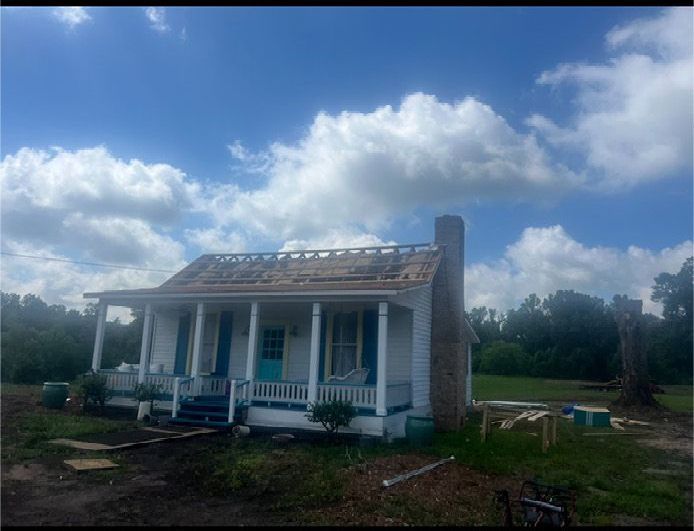 Small white house with blue trim and porch under a cloudy sky, surrounded by grassy yard