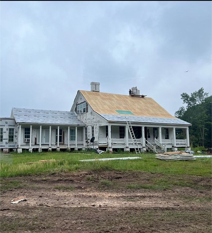 Partially restored white farmhouse with a tan roof on a muddy rural lot under cloudy sky