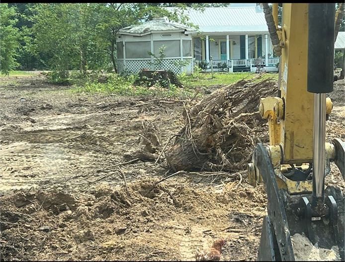 Excavator clearing uprooted tree debris in a muddy yard near a house and porch