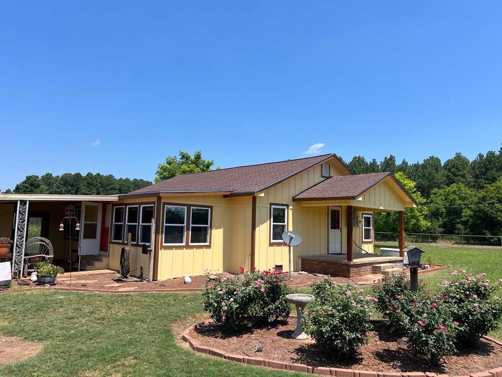 Single-story yellow house with brown roof, front porch, and flower beds under a clear blue sky